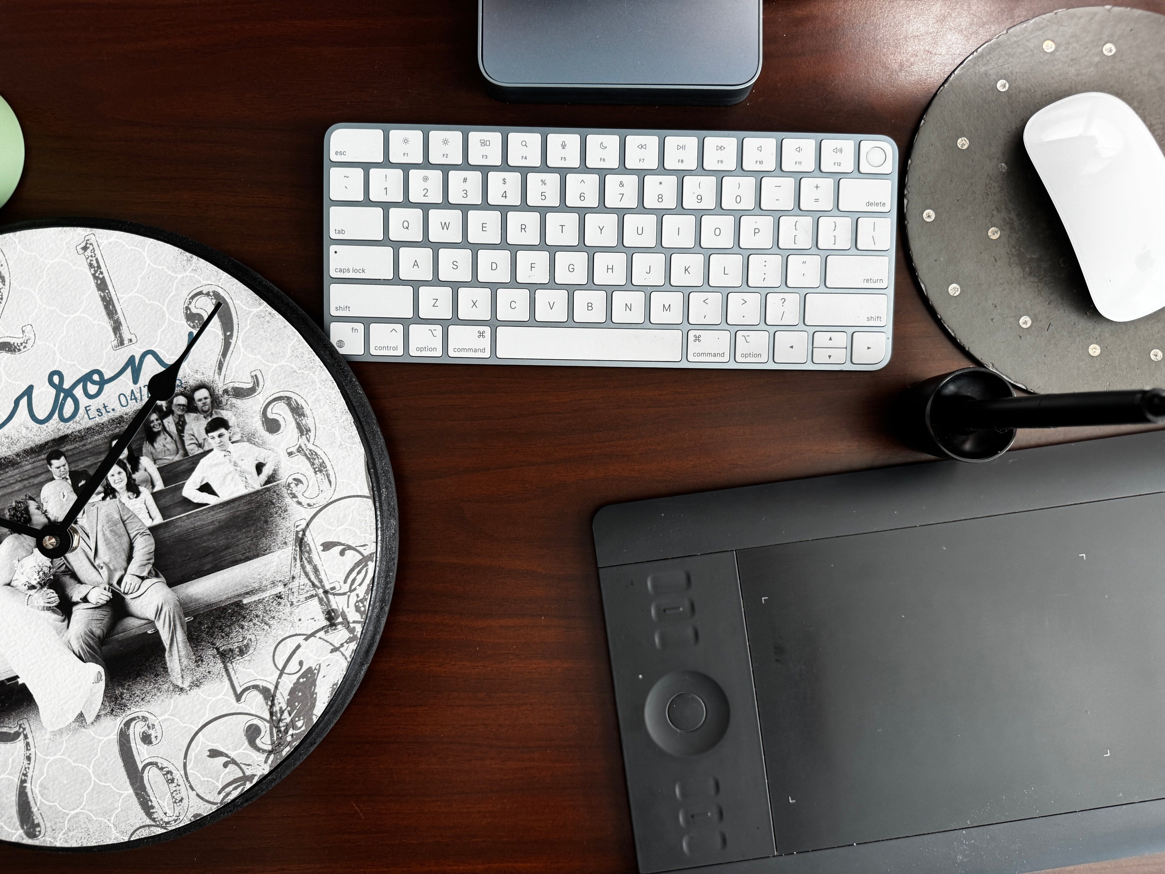 A top-down view of a dark wood desk featuring a white wireless keyboard, a black graphics tablet, a white computer mouse on a gray mousepad, and a round black-and-white illustrated clock.